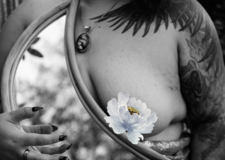 A fat femme person shown standing partly nude and partly wrapped in sequins at a Seattle boudoir session at dusk from the lips to the waist. R's arm and shoulder are tattooed with wings and cats. R is wearing big earrings and a necklace with a large pendant, and is holding a moon-shaped mirror. The image is moody and dark.