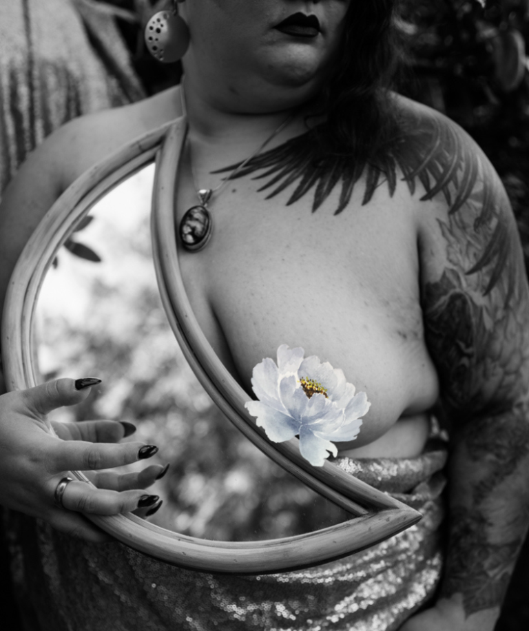 A fat femme person shown standing partly nude and partly wrapped in sequins at a Seattle boudoir session at dusk from the lips to the waist. R's arm and shoulder are tattooed with wings and cats. R is wearing big earrings and a necklace with a large pendant, and is holding a moon-shaped mirror. The image is moody and dark.