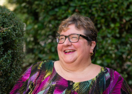 A fat white woman with very short hair, glasses and a colorful top is shown from the chest up at a body positive portrait photo shoot in Seattle. She's laughing and looking toward the edge of the frame, outdoors in front of bushes.
