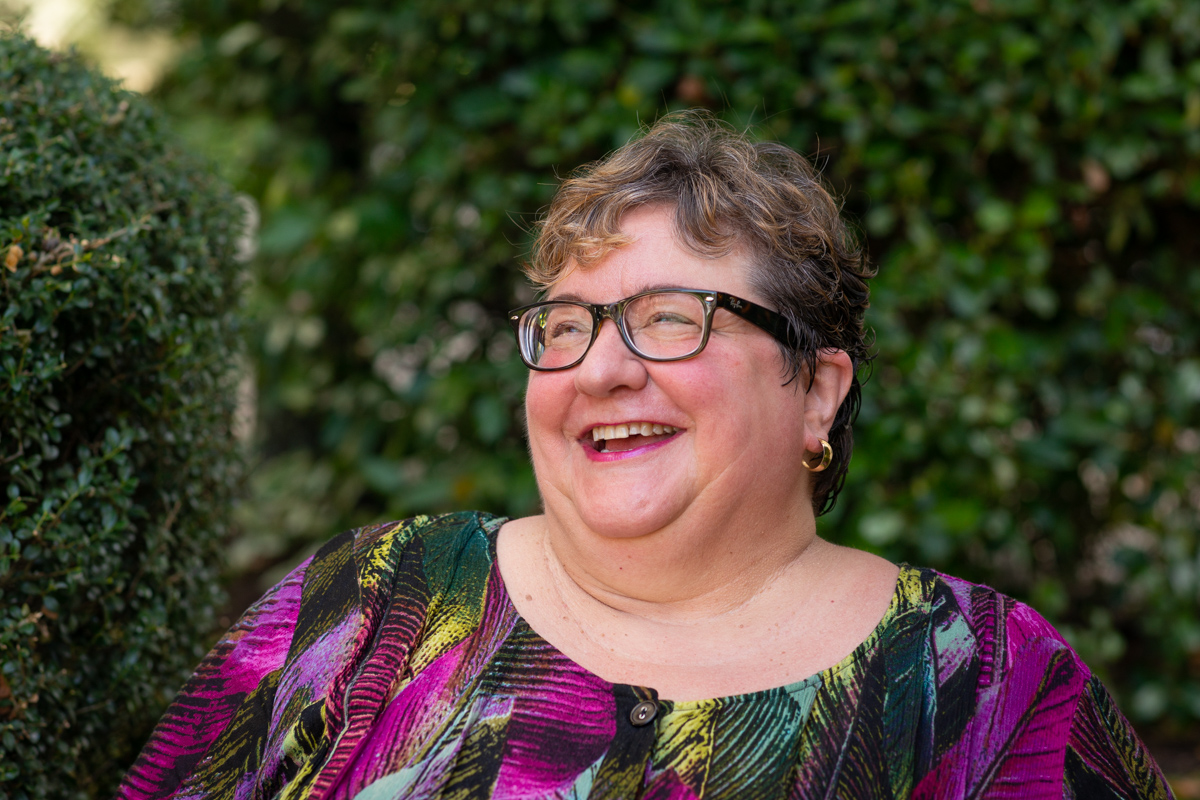 A fat white woman with very short hair, glasses and a colorful top is shown from the chest up at a body positive portrait photo shoot in Seattle. She's laughing and looking toward the edge of the frame, outdoors in front of bushes.