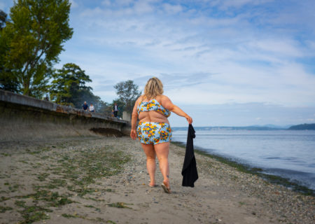 A fat blonde white woman in a colorful two-piece bathing suit discards a black T-shirt as she walks along a sandy beach next to an urban walking path and calm water.