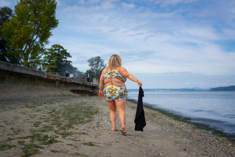 A fat blonde white woman in a colorful two-piece bathing suit discards a black T-shirt as she walks along a sandy beach next to an urban walking path and calm water.