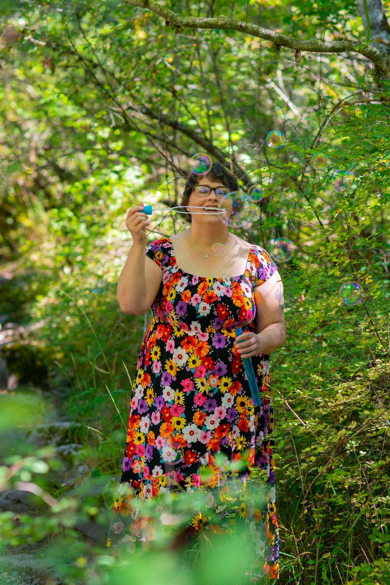 A white woman with short brown hair and glasses blows bubbles in the forest at a body-positive Seattle portrait session in a Washington state park, wearing a floral-print summer dress.