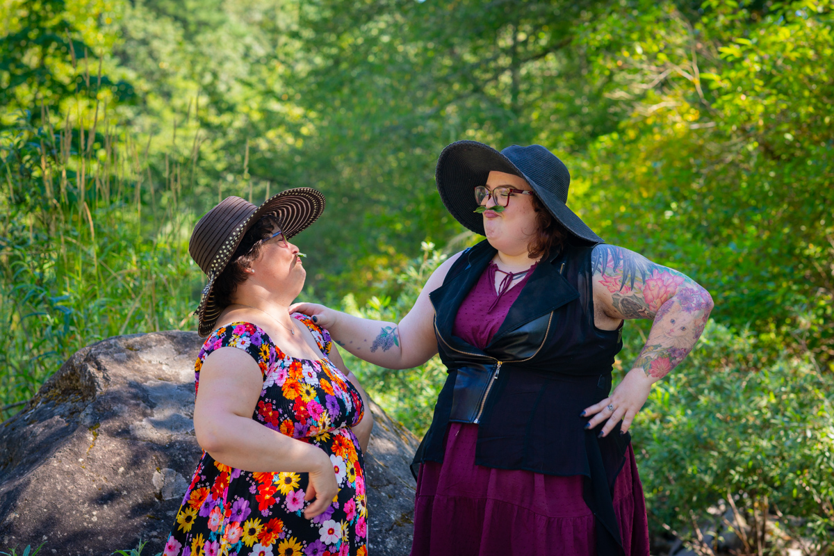 Two larger-bodied white people with short dark hair, glasses and dresses stand outside in front of a forest and boulders. They're playing with leaf moustaches and laughing.