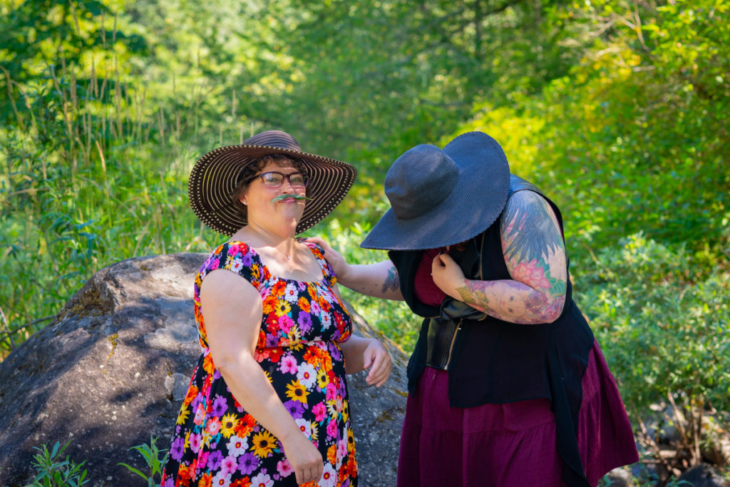 Two larger-bodied white people with short dark hair, glasses and dresses stand outside in front of a forest and boulders. They're playing with leaf moustaches and laughing.