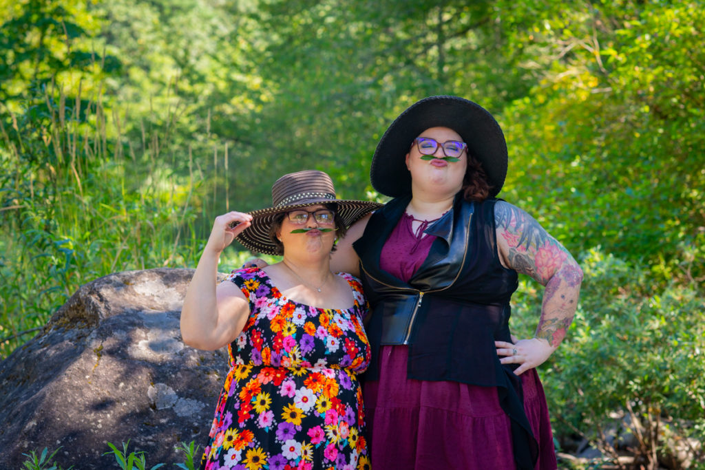 Two larger-bodied white people with short dark hair, glasses and dresses stand outside in front of a forest and boulders. They're playing with leaf moustaches and laughing.