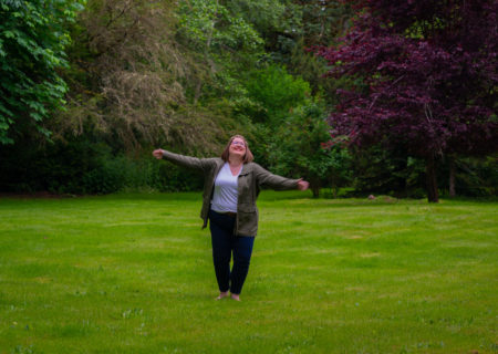 A fat white woman with shoulder-length brown hair throws her arms out and looks up to the sky in a field. She is wearing a white shirt, black pants and a green jacket.
