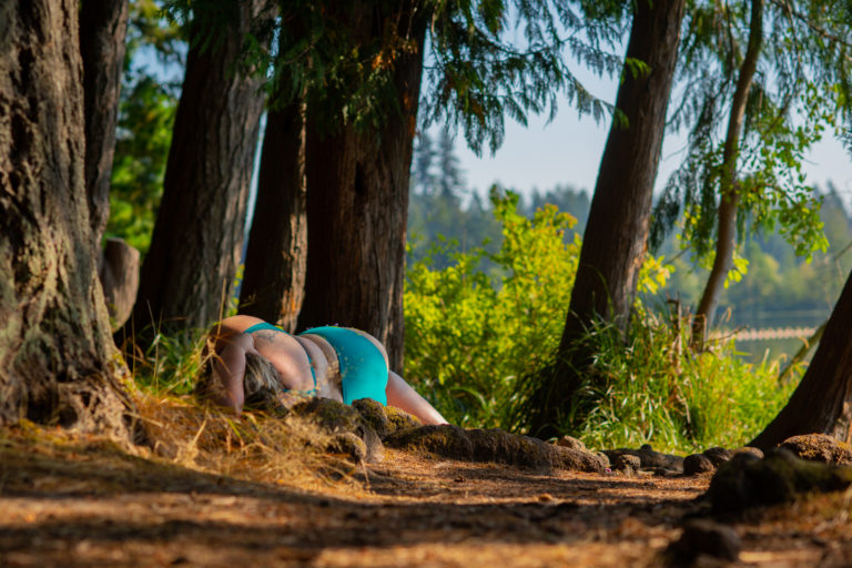 A fat woman in a blue bathing suit lies on the ground in a sun-dappled forest, cradled by tree roots.