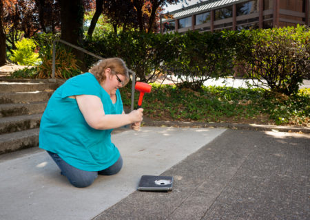 A fat white woman kneels on a sidewalk to smash a bathroom scale using a big orange hammer.