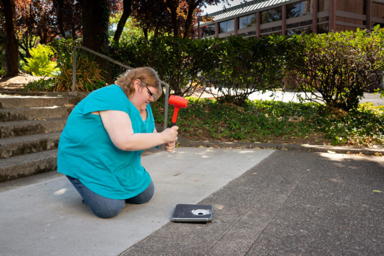 A fat white woman kneels on a sidewalk to smash a bathroom scale using a big orange hammer.