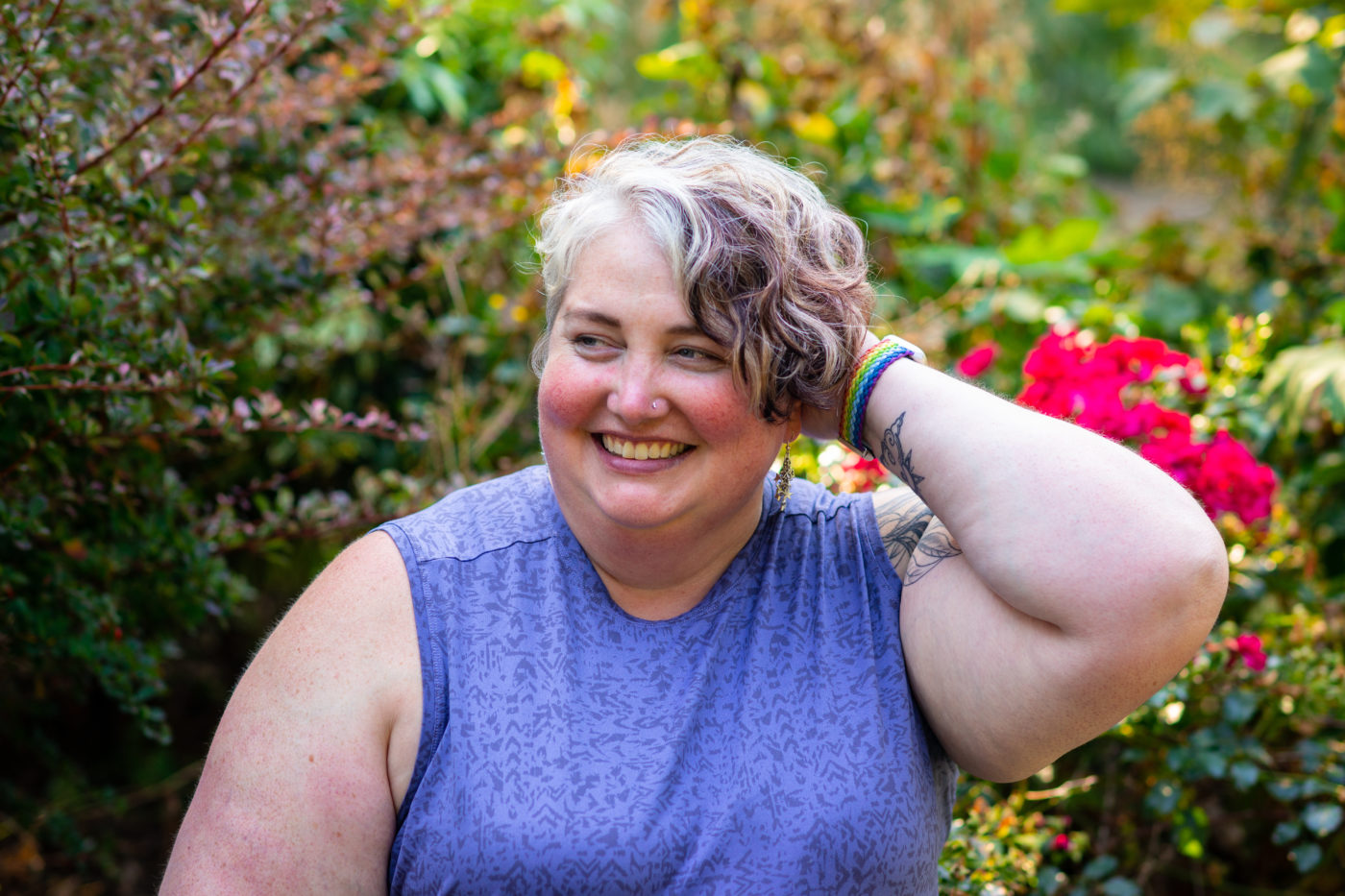 A fat white woman with short brown and gray hair smiles while looking to one side of the camera, with one hand in her hair. She's wearing a sleeveless blue shirt and sitting in front of bushes and bright roses.