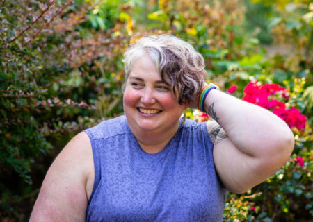 A fat white woman with short brown and gray hair smiles while looking to one side of the camera, with one hand in her hair. She's wearing a sleeveless blue shirt and sitting in front of bushes and bright roses.