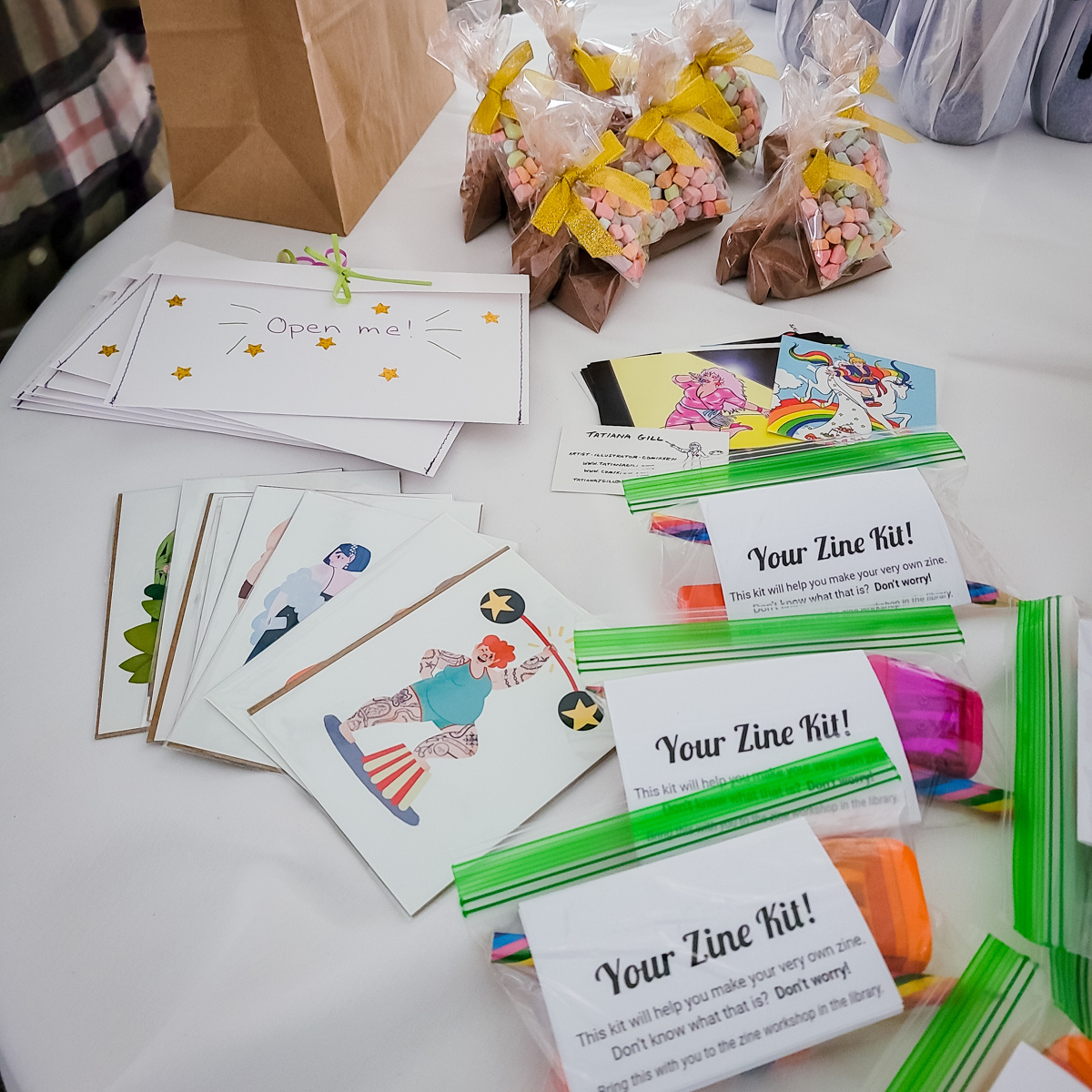 A white table with zine kits in baggies, small fat-positive art prints and stickers, envelopes that say "open me" and little bags of hot chocolate mix with colorful marshmallows.