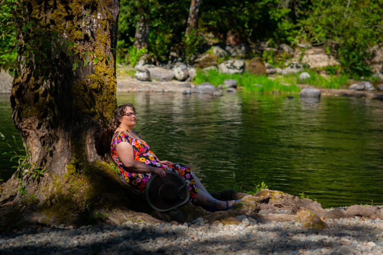 A fat white woman with short brown hair sits against a tree trunk by a river in the sun, holding a wide-brimmed hat and smiling gently.