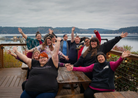 A group of fat folks smiling and joyfully waving their arms in the air. They're sitting or standing around a picnic table in front of calm water, boats and low hills in the distance under a gray sky.