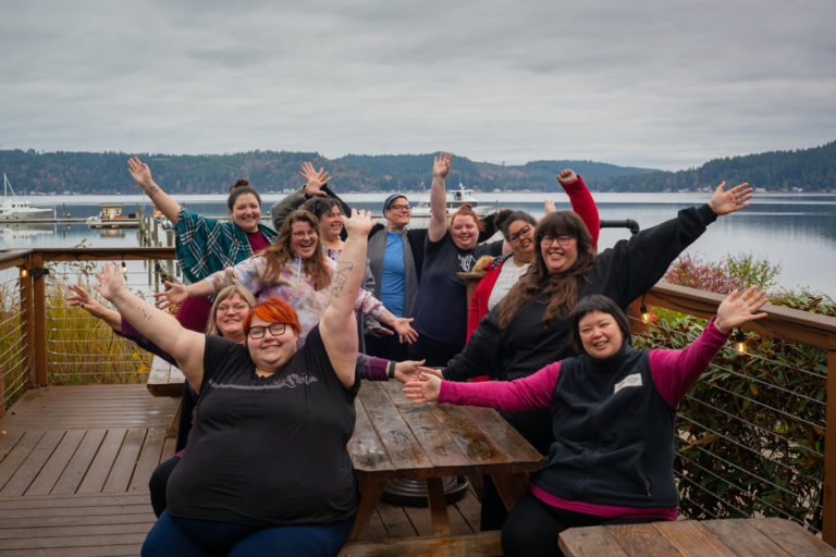 A group of fat folks smiling and joyfully waving their arms in the air. They're sitting or standing around a picnic table in front of calm water, boats and low hills in the distance under a gray sky.