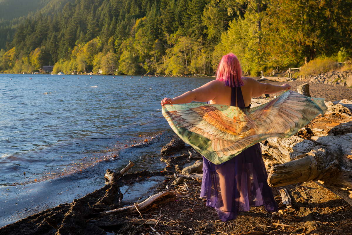[Image: A fat white woman with pink hair stands on the shore of a wooded lake in a purple sleeveless dress, with a wing-print scarf blowing in the wing behind her outstretched arms.]