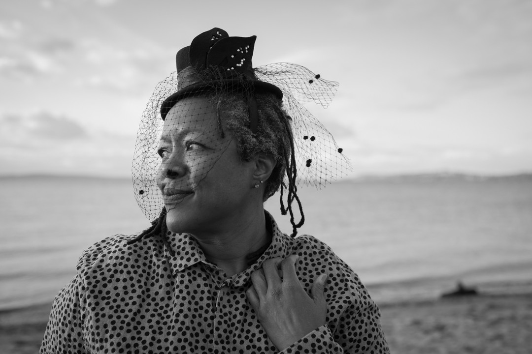 A Black plus-size woman in a dotted button-down shirt and a little top hat with a veil looks to one side, smiling slightly, on a beach.