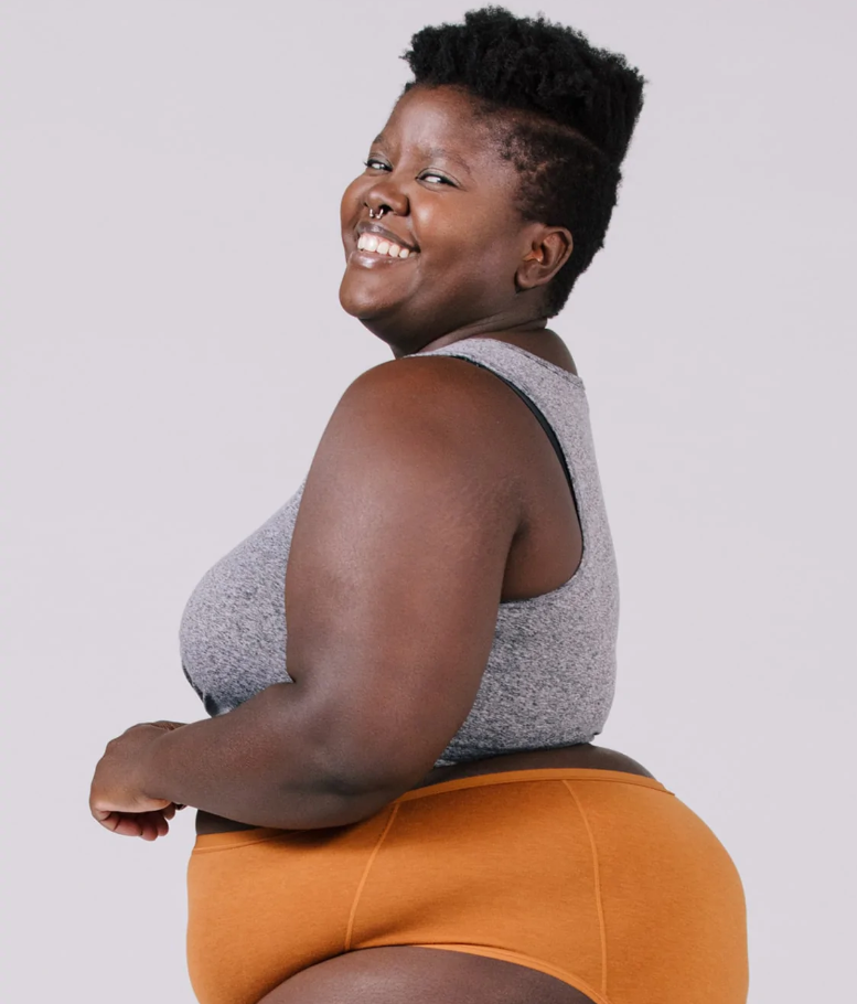 A fat black person wearing a gray tank top and orange underwear smiles at the camera