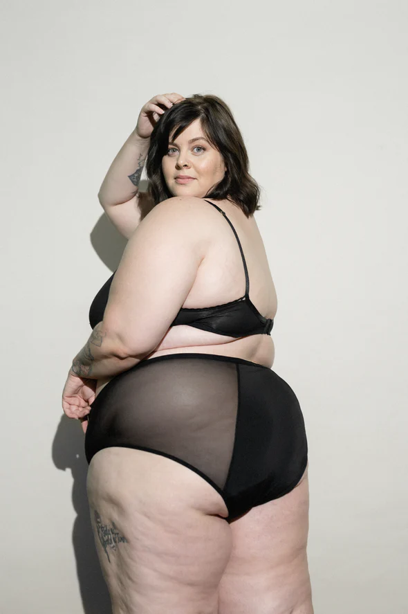 A fat white person wearing black underwear looks at the camera over their shoulder