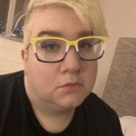 A fat white non-binary person with light blonde hair and shaved sides, wearing non-binary flag glasses frames