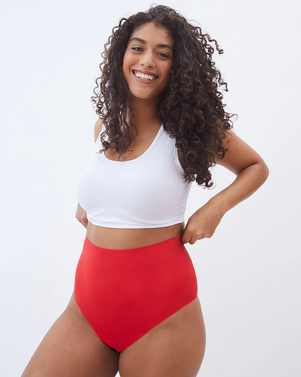 A thin person with curly hair wearing a white bra and orange underwear smiles at the camera while holding the waistband of their underwear