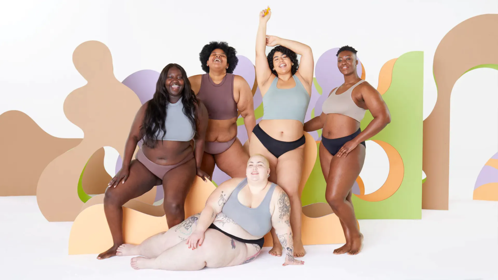 A group of people of various sizes wearing different bras and underwear pose in front of an abstract background smiling at the camera