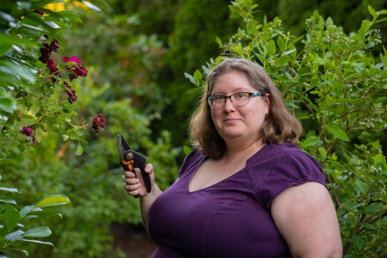 Lindley, a fat white woman with shoulder-length hair and glasses, stands in a garden wearing a dress and holding a pair of garden clippers. She's smiling mischievously.