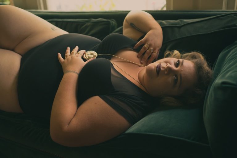 A fat feminine person lies on their back on a velvet couch, wearing a black leotard and a gold necklace with a fat body pendant. They're looking pensively at the camera.