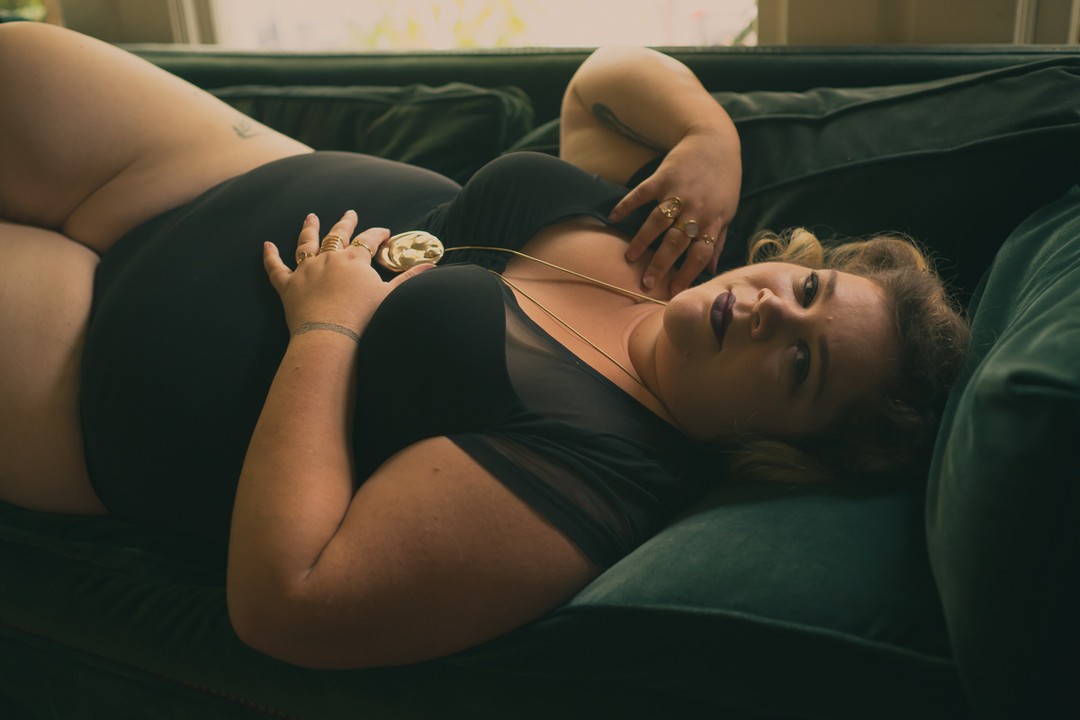 A fat feminine person lies on their back on a velvet couch, wearing a black leotard and a gold necklace with a fat body pendant. They're looking pensively at the camera.