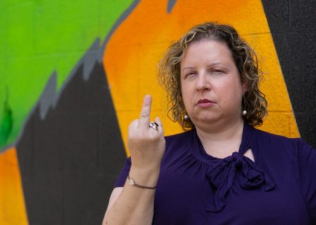 A white woman narrows her eyes and sticks her middle finger up at the camera in front of a colorful mural.