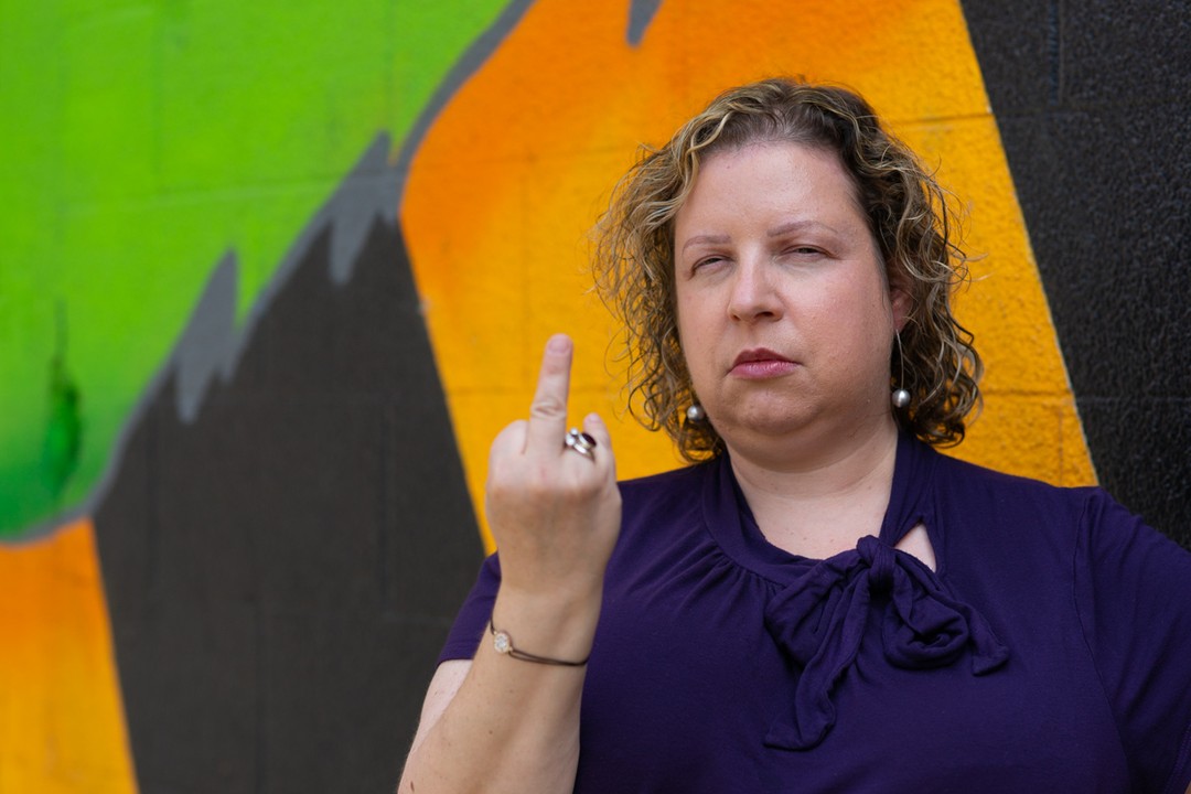 A white woman narrows her eyes and sticks her middle finger up at the camera in front of a colorful mural.
