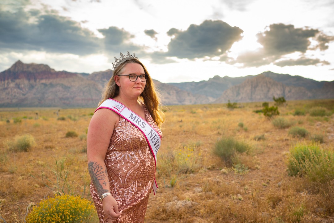 A white woman with long hair, tattoos, a sparkly dress, Mrs. Nevada sash and crown stands in the desert with distant mountains and bright sky. She's at a Body Liberation Photos fat-positive portrait session.