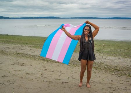 A Black trans woman in a bathing suit stands on a cloudy beach and smiles while holding up a trans pride flag.