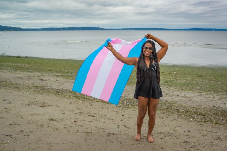 A Black trans woman in a bathing suit stands on a cloudy beach and smiles while holding up a trans pride flag.