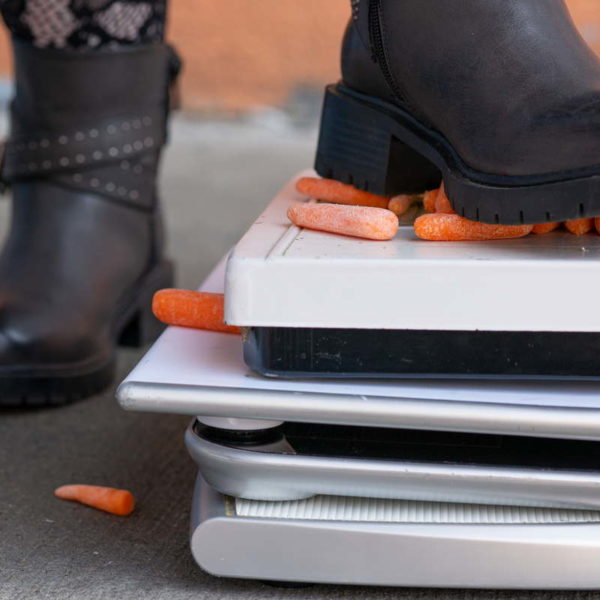 A woman wearing black boots stomps on a pile of bathroom scales laden with baby carrots.