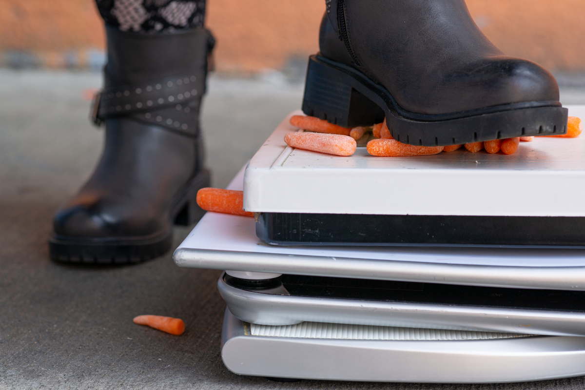 A woman wearing black boots stomps on a pile of bathroom scales laden with baby carrots.