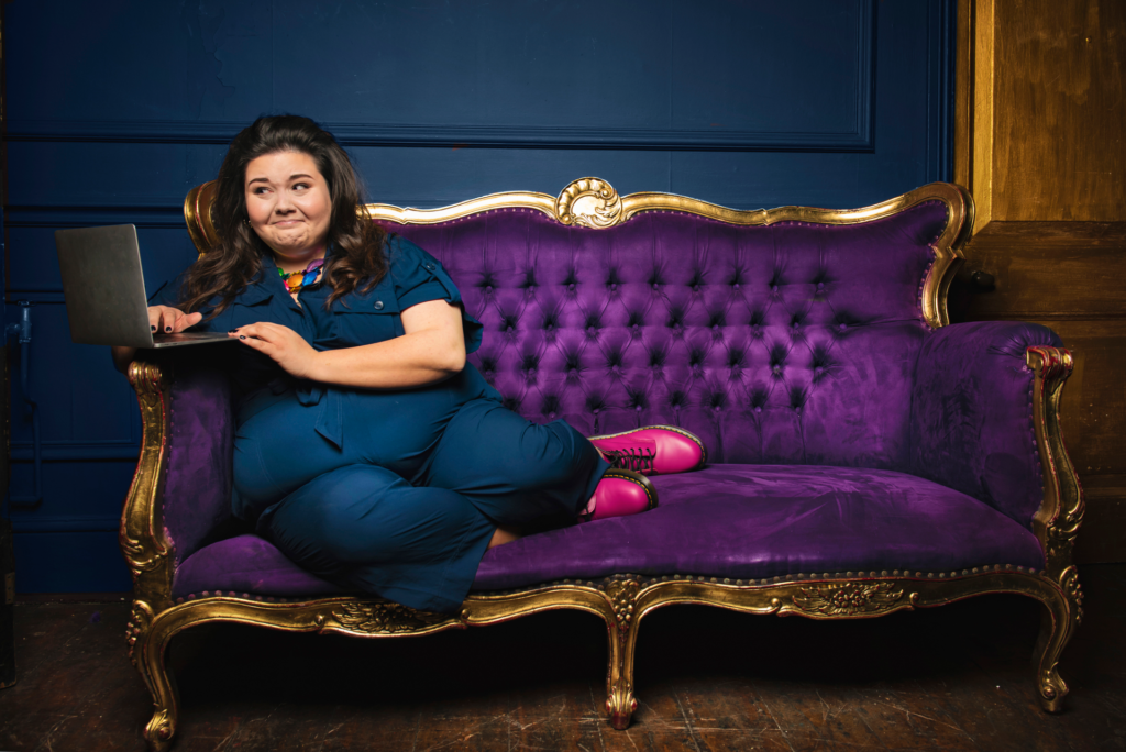 A fat person sits on a purple couch with a gold frame looking amusedly at their laptop