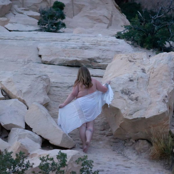 In this fine art photograph by Lindley Ashline, a plus-size white woman clad in only the white scarf she holds wanders through a rocky landscape at dusk.