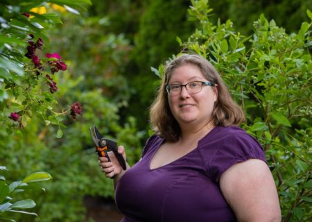 Lindley, a fat white woman, stands in a garden holding clippers, wearing a purple dress and looking at the camera with an interested expression.