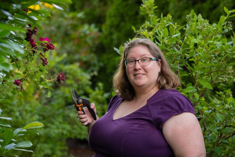 Lindley, a fat white woman, stands in a garden holding clippers, wearing a purple dress and looking at the camera with an interested expression.