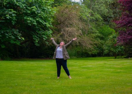 A fat white woman in casual clothing throws her arms up to the sky joyfully on a grassy lawn with trees in the background.
