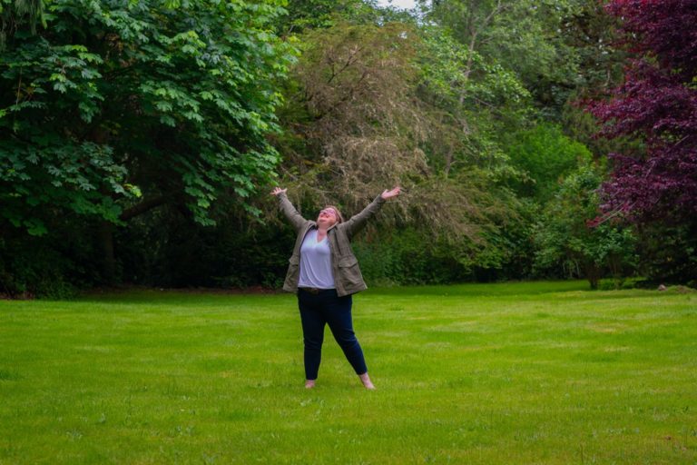 A fat white woman in casual clothing throws her arms up to the sky joyfully on a grassy lawn with trees in the background.