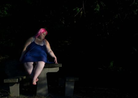 Danielle Bex, a fat white non-binary femme in a purple summer dress and bright pink hair, sits on a picnic table in near-darkness and traces a design on the table with a finger.
