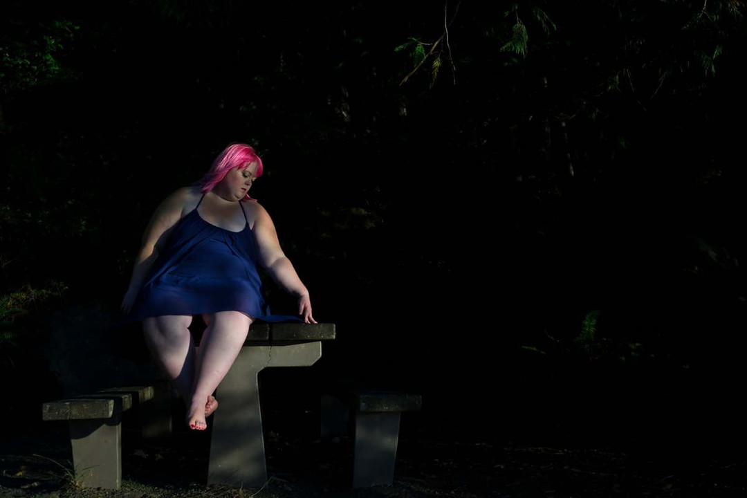 Danielle Bex, a fat white non-binary femme in a purple summer dress and bright pink hair, sits on a picnic table in near-darkness and traces a design on the table with a finger.