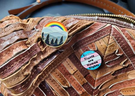 A close up of a brown leather purse with two enamel pins. One has trees, mountains and a rainbow; the other has the trans flag with the words "trans rights are human rights" on top.