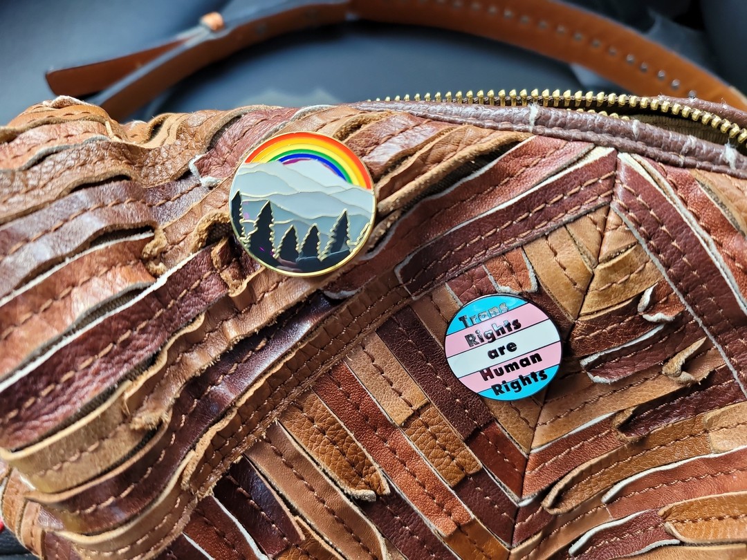 A close up of a brown leather purse with two enamel pins. One has trees, mountains and a rainbow; the other has the trans flag with the words "trans rights are human rights" on top.