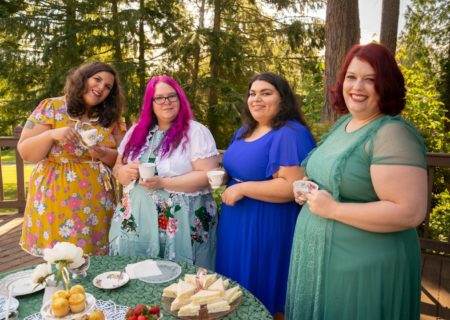 Four smiling fat people, all feminine in dresses with medium to long hair, standing at a tea table on a suburban deck and holding tea cups.