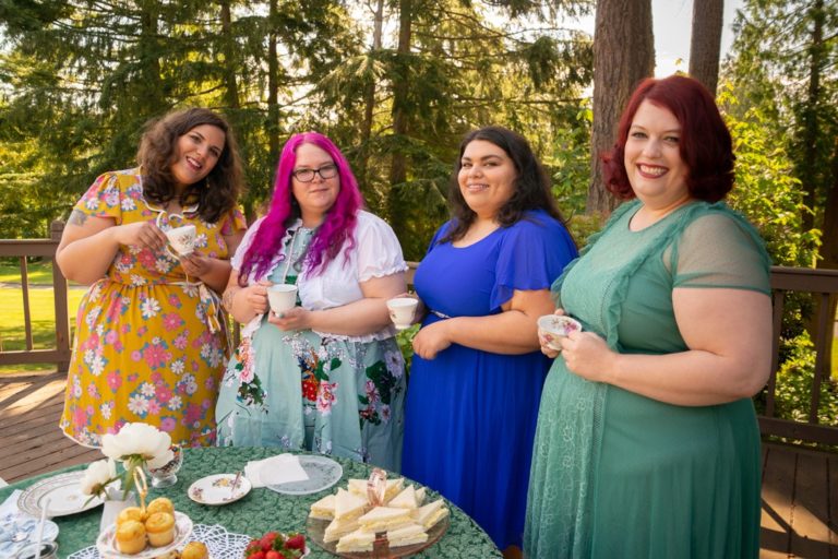 Four smiling fat people, all feminine in dresses with medium to long hair, standing at a tea table on a suburban deck and holding tea cups.