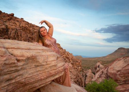 A plus-size white woman in a pink sequined gown with long dark blonde hair arches an arm and looks off to the distance while leaning on a boulder in a desert landscape.